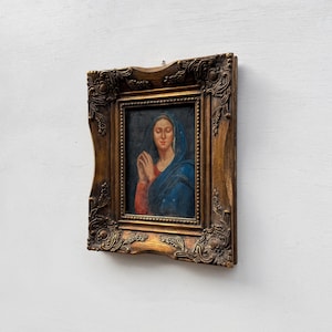 Könnte beinhalten: Gerahmtes religiöses Gemälde mit einer Frau in blauem Kopftuch und roten Ärmeln, die Hände zum Gebet gefaltet. Der verzierte, goldfarbene Rahmen verleiht dem Kunstwerk einen klassischen Touch.