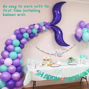 May include: A mermaid-themed birthday party setup with a balloon arch in shades of purple, teal, and white. A large purple mermaid tail balloon is attached to the wall. The table is decorated with a "Happy Birthday" banner and a cake.