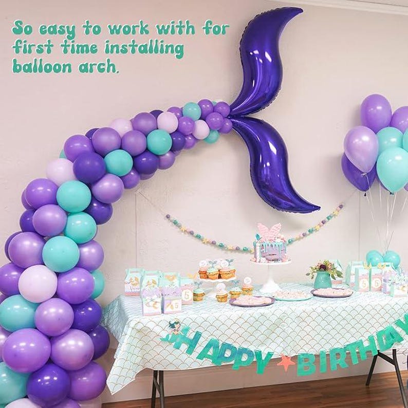 May include: A mermaid-themed birthday party setup with a balloon arch in shades of purple, teal, and white. A large purple mermaid tail balloon is attached to the wall. The table is decorated with a "Happy Birthday" banner and a cake.