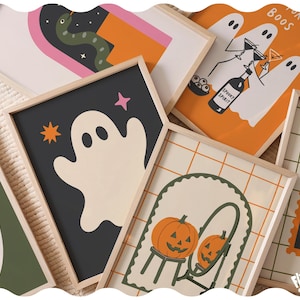 May include: A collection of framed art prints featuring Halloween-themed illustrations. Designs include ghosts, a snake, a pumpkin in a mirror, and text reading FOR THE BOOS, SPOOKY SPIRIT, and FIRUM MARKET. The prints are in light wooden frames.