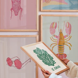 May include: A collection of framed art prints with a beach theme. Prints include a pink swimsuit, a lobster, sunglasses, and a green coral design with the text "LE CORAIL". The frames are light wood.