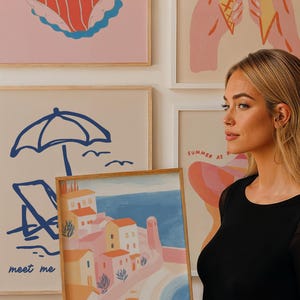 May include: A framed art print featuring a coastal town scene with buildings in shades of pink, orange, and white, set against a blue sea and sky. Other prints in the background depict summer-themed illustrations, including a swimsuit, ice cream cones, and beach elements.