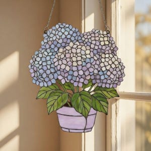 Könnte beinhalten: Buntglas-Fensteraufhänger mit einer Hortensienpflanze in einem lavendelfarbenen Topf. Die Blüten sind eine Mischung aus Blau, Lila und Weiß, mit grünen Blättern. Aufgehängt an einer silbernen Kette, ideal zur Dekoration.
