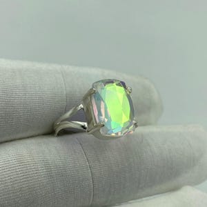 May include: A silver ring featuring a large, rectangular iridescent gemstone. The gemstone displays a spectrum of colors, including green, yellow, and pink, and is set in a split-shank band. The ring is shown against a white background.