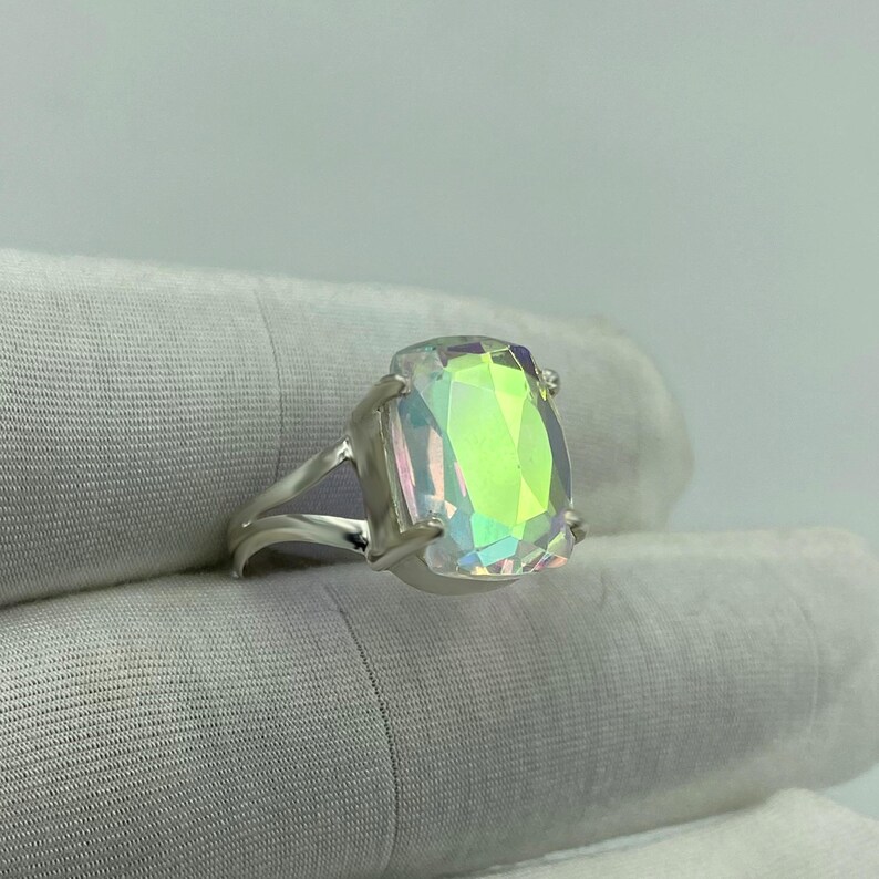 May include: A silver ring featuring a large, rectangular iridescent gemstone. The gemstone displays a spectrum of colors, including green, yellow, and pink, and is set in a split-shank band. The ring is shown against a white background.