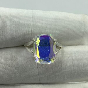 May include: A silver ring featuring a large, rectangular, iridescent gemstone. The gemstone displays a spectrum of colors, including blue, yellow, and purple, creating a shimmering effect. The ring's band is a simple, split-shank design.