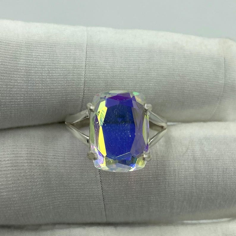 May include: A silver ring featuring a large, rectangular, iridescent gemstone. The gemstone displays a spectrum of colors, including blue, yellow, and purple, creating a shimmering effect. The ring's band is a simple, split-shank design.