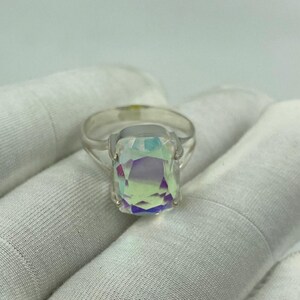 May include: A silver ring featuring a large, rectangular iridescent gemstone. The gemstone displays a spectrum of colors, including purple, green, and yellow. The ring is set on a white background.