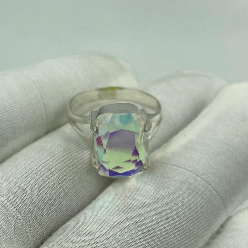 May include: A silver ring featuring a large, rectangular iridescent gemstone. The gemstone displays a spectrum of colors, including purple, green, and yellow. The ring is set on a white background.