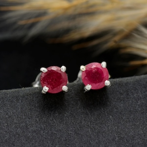 May include: A pair of stud earrings featuring round, deep red gemstones set in silver. The gemstones are the focal point, with a polished surface and a vibrant color. The earrings are displayed on a dark, textured surface.