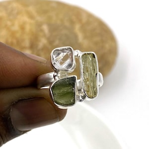 Puede incluir: Un anillo de plata con tres piedras preciosas de forma irregular. Las piedras son transparentes, verde claro y verde oscuro. El anillo está sobre un fondo blanco.