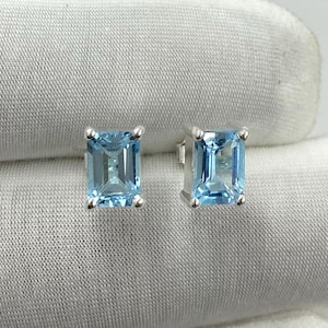 May include: A pair of silver stud earrings, each with a rectangular, light blue gemstone. The gemstones are set in a four-prong setting. The earrings are presented on a white fabric background. A classic jewellery item.