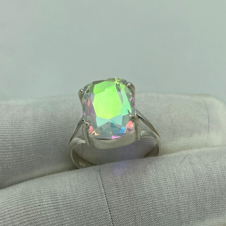 May include: A silver ring featuring a large, iridescent, rectangular gemstone. The gemstone displays a spectrum of colors, including green, yellow, and blue, reflecting light. The ring's band is a simple, polished silver design, showcasing the prominent stone.