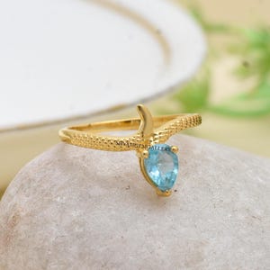 May include: A gold-colored ring with a textured snake design. The ring features a pear-shaped, light blue gemstone. The ring is displayed on a light-colored, textured surface.