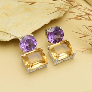 Puede incluir: Pendientes plateados con una amatista morada redonda sobre una citrina amarilla rectangular. Las joyas se colocan sobre un fondo neutro con una piedra clara y hierba seca.