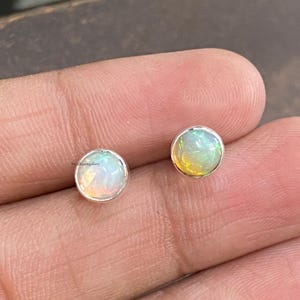 May include: A pair of round opal stud earrings. Each earring features a vibrant, iridescent opal stone set in a silver bezel. The opals display a mix of colors, including white, yellow, and green. The earrings are small and delicate.