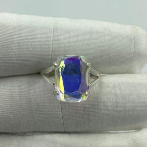 May include: A silver ring featuring a large, rectangular iridescent gemstone. The gemstone displays a spectrum of colors, including blue, yellow, and purple. The ring's band is a simple, elegant design.