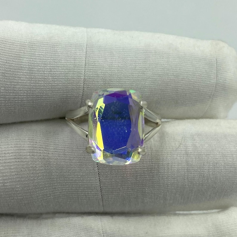 May include: A silver ring featuring a large, rectangular iridescent gemstone. The gemstone displays a spectrum of colors, including blue, yellow, and purple. The ring's band is a simple, elegant design.