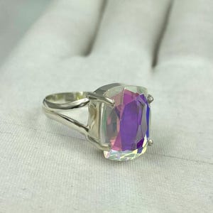 May include: A silver ring featuring a large, rectangular iridescent gemstone. The gemstone displays a spectrum of colors, including purple, pink, and blue. The ring's band has a split design, adding a modern touch to the jewelry.