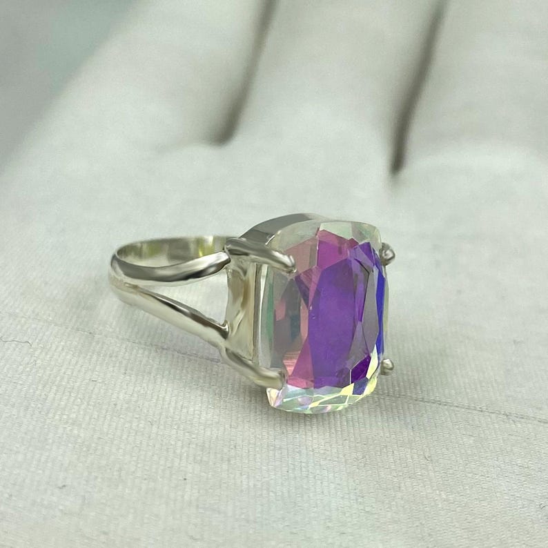 May include: A silver ring featuring a large, rectangular iridescent gemstone. The gemstone displays a spectrum of colors, including purple, pink, and blue. The ring's band has a split design, adding a modern touch to the jewelry.