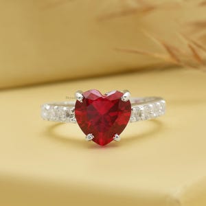 Puede incluir: Un anillo de plata con una gran gema roja en forma de corazón. La banda está adornada con gemas claras más pequeñas. El diseño del anillo se presenta sobre un fondo amarillo suave, resaltando el rojo vibrante de la piedra central.
