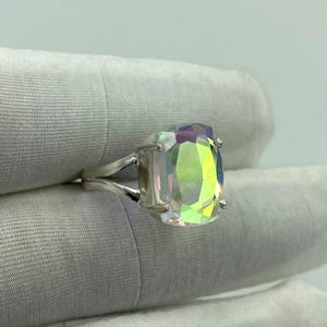 May include: A silver ring featuring a large, iridescent, rectangular-cut gemstone. The stone displays a spectrum of colors, including green, yellow, and purple. The ring's band splits into two strands on each side, creating a unique design.