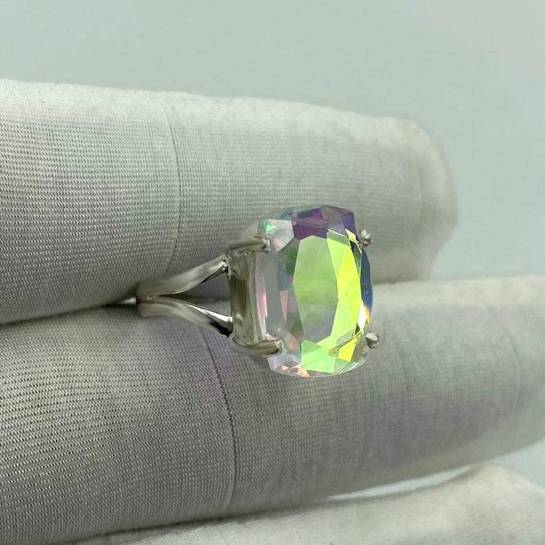 May include: A silver ring featuring a large, iridescent, rectangular-cut gemstone. The stone displays a spectrum of colors, including green, yellow, and purple. The ring's band splits into two strands on each side, creating a unique design.