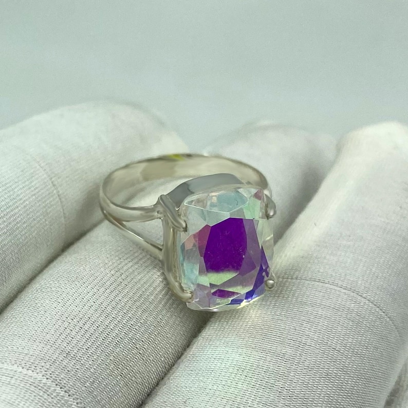 May include: A silver ring featuring a large, rectangular, iridescent gemstone. The gemstone displays a spectrum of colors, including purple, blue, and green, and is set in a simple, elegant silver band. The ring is displayed on a white surface.