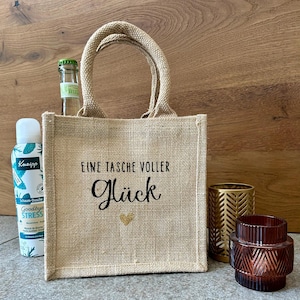 Könnte beinhalten: Eine kleine Jutetasche mit dem Text "EINE TASCHE VOLLER Glück" und einem goldenen Herz. Neben der Tasche befinden sich eine Flasche Kneipp Goodbye Stress Duschschaum, eine kleine Glasflasche und zwei dekorative Kerzenhalter.