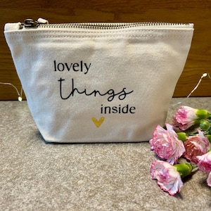 Könnte beinhalten: Ein cremefarbener Canvas-Beutel mit Reißverschluss. Auf der Vorderseite ist der Text "lovely things inside" mit einem gelben Herz aufgedruckt. Rechts vom Beutel befinden sich rosa und weiße Blumen.