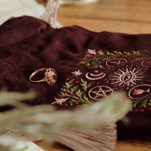 May include: A gold ring with a cluster of red gemstones sits on a dark burgundy velvet pouch. The pouch is embroidered with celestial symbols and green and red accents. The ring is the focal point of the image.