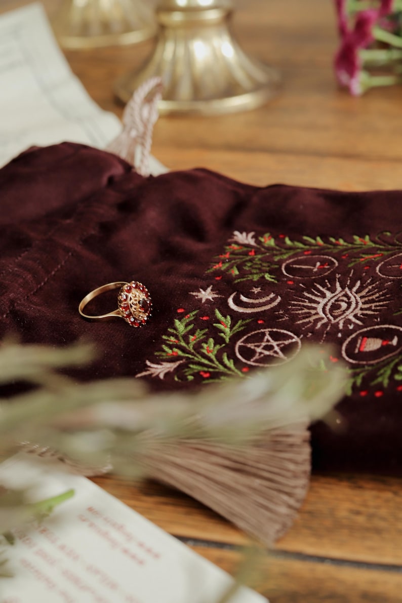 May include: A gold ring with a cluster of red gemstones sits on a dark burgundy velvet pouch. The pouch is embroidered with celestial symbols and green and red accents. The ring is the focal point of the image.