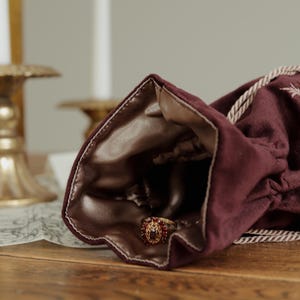 May include: A burgundy velvet drawstring bag with a gold satin lining, holding a gold ring with red gemstones. The ring is in focus, with the bag partially open. The background includes candles and a wooden surface.