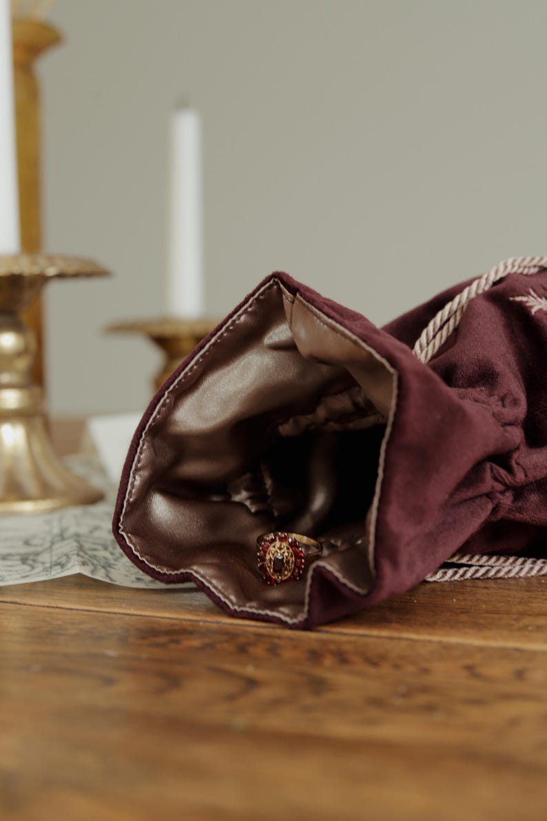 May include: A burgundy velvet drawstring bag with a gold satin lining, holding a gold ring with red gemstones. The ring is in focus, with the bag partially open. The background includes candles and a wooden surface.