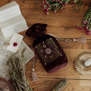 May include: A burgundy velvet drawstring bag with an embroidered celestial design featuring a crescent moon, sun, and other symbols. The bag is on a wooden surface with dried herbs, flowers, and a map.