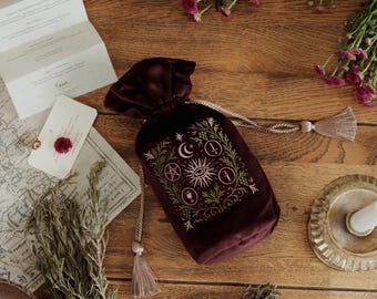 Puede incluir: Una bolsa de terciopelo burdeos con cordón y diseño celestial bordado con luna creciente, sol y otros símbolos. La bolsa está sobre una superficie de madera con hierbas secas, flores y un mapa.