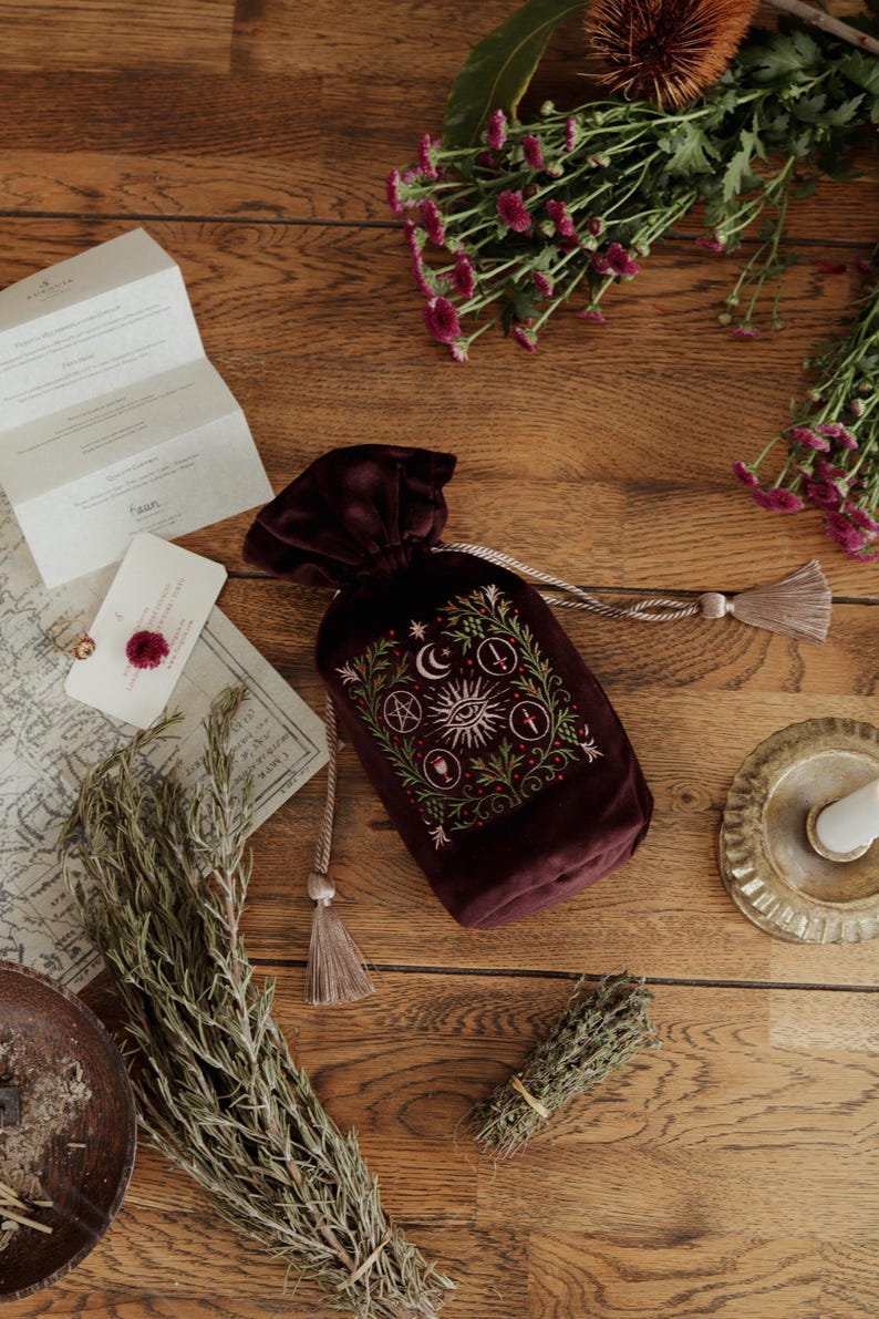 May include: A burgundy velvet drawstring bag with an embroidered celestial design featuring a crescent moon, sun, and other symbols. The bag is on a wooden surface with dried herbs, flowers, and a map.