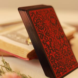 Pode incluir: Um baralho de cartas de tarô com borda preta e design vermelho ornamentado. As cartas estão anguladas, revelando os intrincados padrões e símbolos. O baralho está sobre uma superfície branca com um livro ao fundo.
