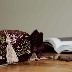 May include: A dark burgundy velvet drawstring bag with embroidered celestial and botanical designs in white and green. The bag features a long, beige tassel and a matching velvet bow closure. The bag is on a wooden surface.