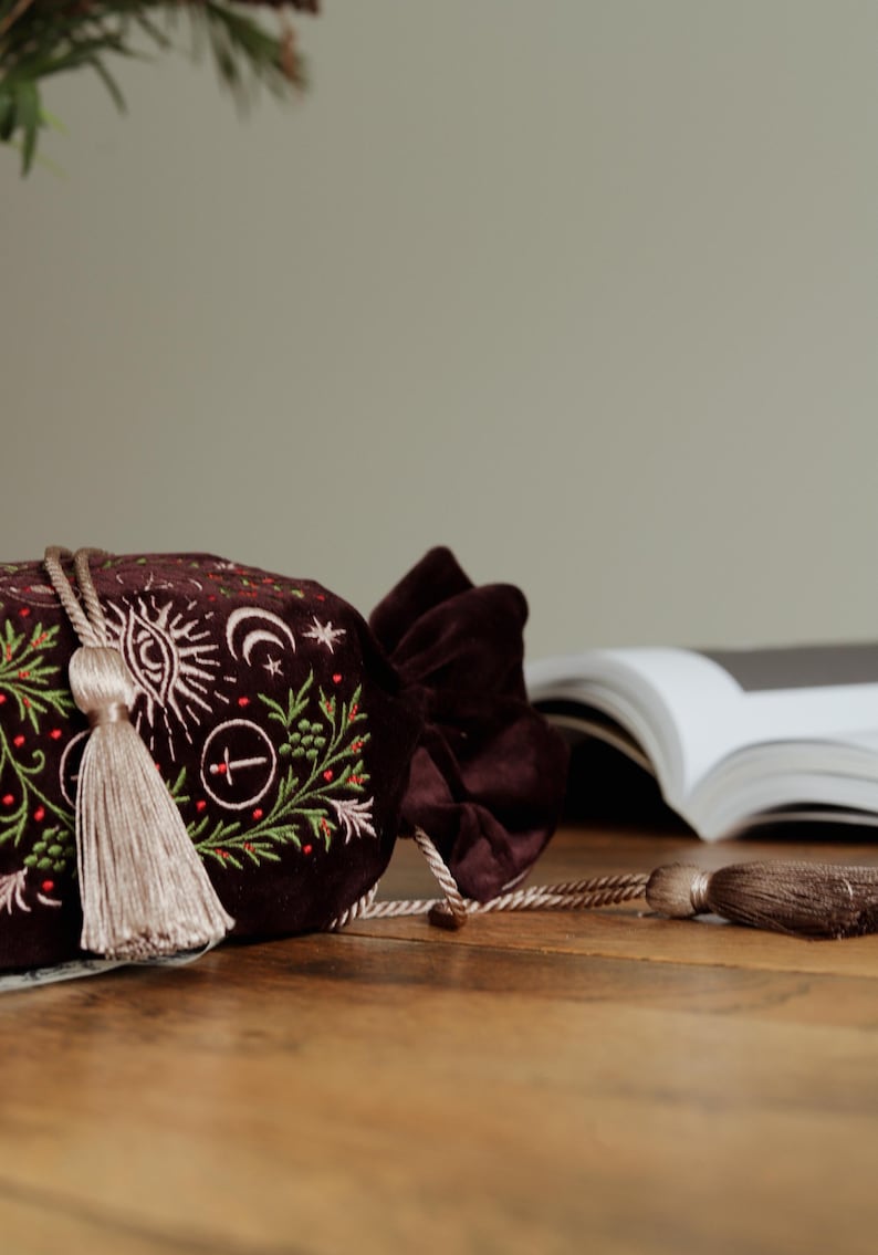 May include: A dark burgundy velvet drawstring bag with embroidered celestial and botanical designs in white and green. The bag features a long, beige tassel and a matching velvet bow closure. The bag is on a wooden surface.