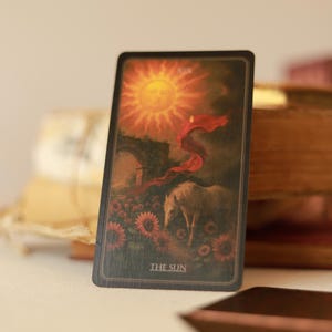 May include: A tarot card featuring a radiant sun with a face, a white horse, and sunflowers. The card is labeled "THE SUN" at the bottom. The card is set against a backdrop of a bridge and a flowing red cloth.
