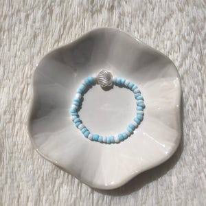 May include: A bracelet made of light blue and white beads, with a silver seashell charm. The bracelet is presented on a white, scalloped-edge dish. The beads are small, and the overall design is delicate.