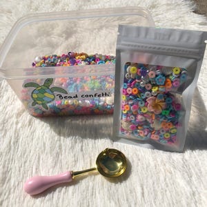 May include: A clear plastic container and a sealed bag filled with colourful bead confetti, alongside a pink-handled, gold-coloured wax melting spoon. The container has a turtle graphic and the words "Bead confetti".