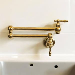 Puede incluir: Un grifo de llenado de ollas dorado, montado en la pared, con un brazo de doble articulación. El grifo está instalado sobre un fregadero blanco con moldura de madera. El diseño es clásico y elegante, adecuado para una cocina.