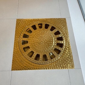 May include: A square, hammered brass floor drain cover with a circular, slotted centre. The drain cover is a bright gold colour and sits on a white tiled floor. The circular centre has evenly spaced slots.