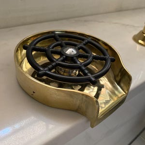 K&ouml;nnte beinhalten: Ein Messing-Aschenbecher mit einem schwarzen Steuerraddesign. Der Aschenbecher hat eine gebogene Form mit einer kleinen Lippe auf einer Seite. Das Steuerrad ist in der Mitte des Aschenbechers.