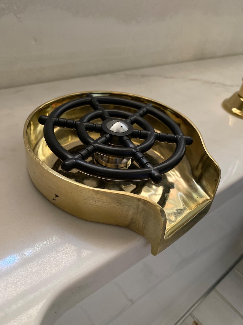 K&ouml;nnte beinhalten: Ein Messing-Aschenbecher mit einem schwarzen Steuerraddesign. Der Aschenbecher hat eine gebogene Form mit einer kleinen Lippe auf einer Seite. Das Steuerrad ist in der Mitte des Aschenbechers.