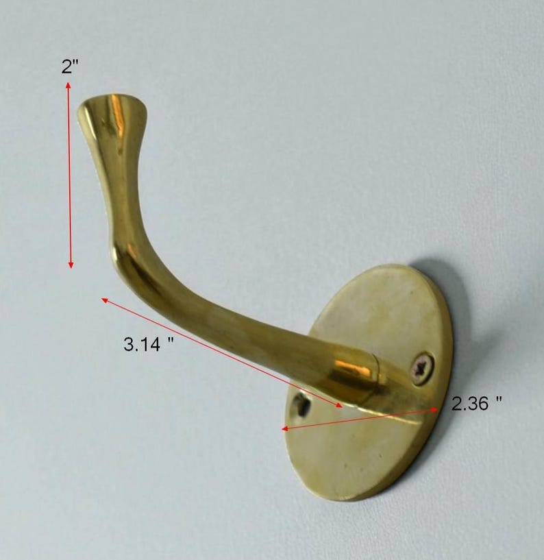 May include: A brass wall hook with an oval base and curved arm. The hook is 2 inches tall and 3.14 inches long, with a base that is 2.36 inches wide. The hook is mounted on a white wall.
