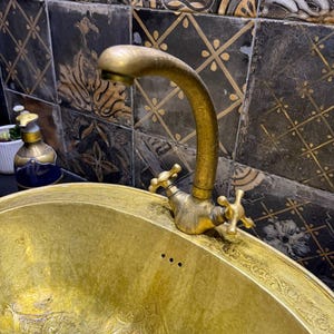 May include: A vintage brass sink with an ornate design and matching faucet. The sink has a curved spout and cross-shaped handles. The sink is gold in color and has an etched pattern around the rim. The background features decorative tiles.