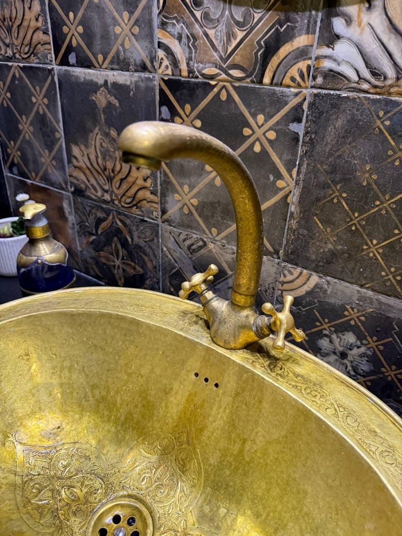May include: A vintage brass sink with an ornate design and matching faucet. The sink has a curved spout and cross-shaped handles. The sink is gold in color and has an etched pattern around the rim. The background features decorative tiles.